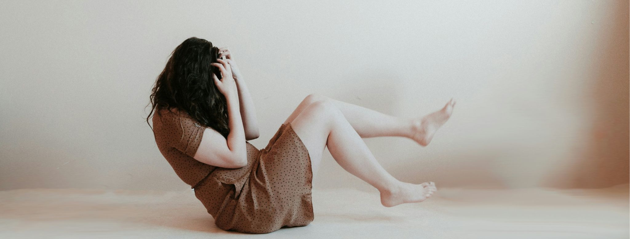 An anxious woman sits on the floor in a brightly lit room, her body language reflecting tension and unease. She clasps her hands tightly, shoulders hunched, as sunlight streams in, creating a stark contrast with the emotional struggle evident on her face. The room's brightness serves as a poignant backdrop to the internal turmoil she experiences, emphasizing the vulnerability of the moment